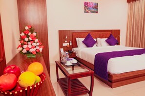 A hotel room with a wooden bed, a nightstand, a glass coffee table, a bouquet of flowers, and a fruit basket. - Aloka Stay Inn (Vellore)