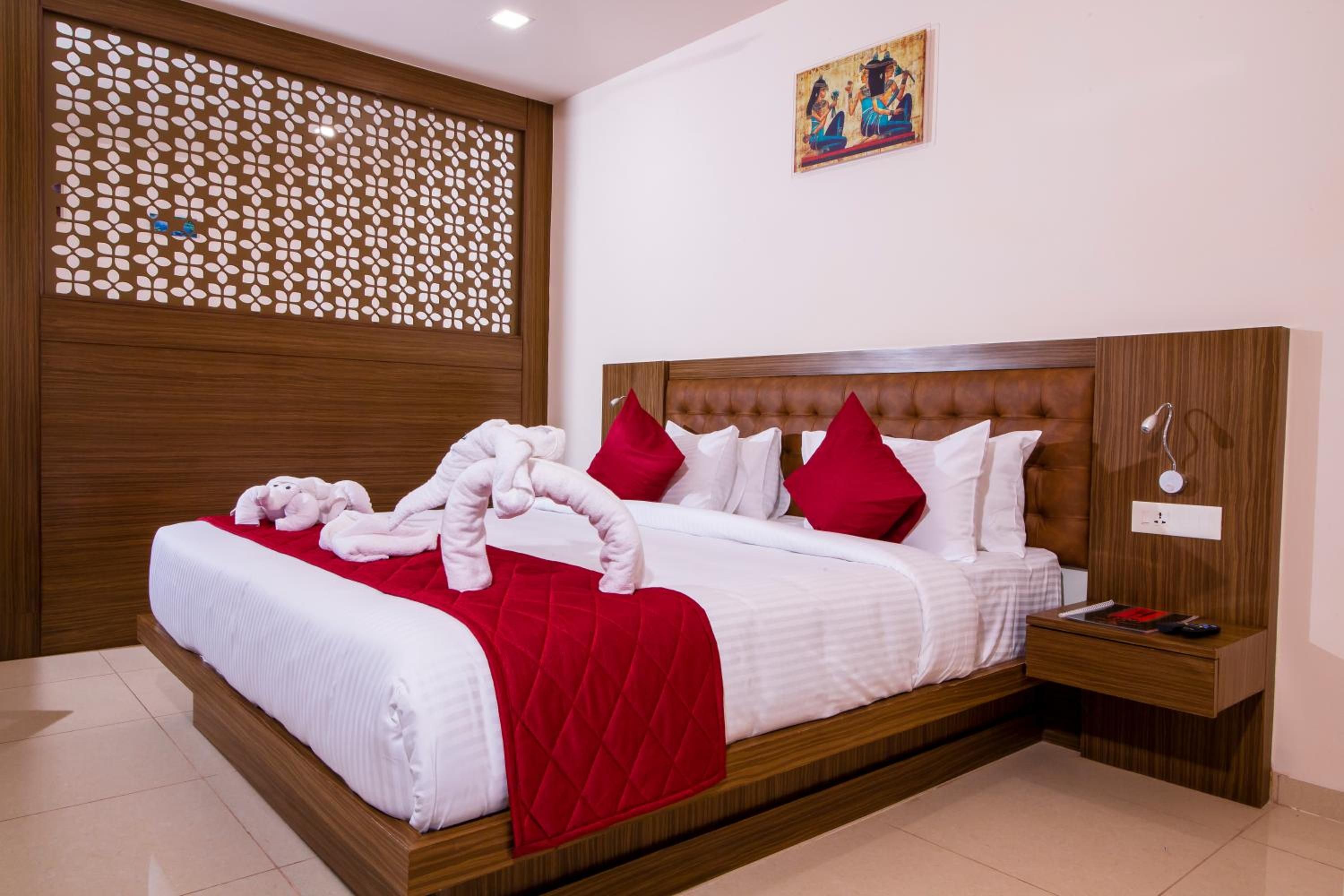 A hotel room with a wooden headboard, a bed with white and red bedding, a decorative wall panel, and a painting on the wall.