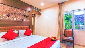 Habitación de hotel con una cama grande, almohadas rojas, colcha roja, cabecero de madera, ventana con vistas a la vegetación y una silla con un frutero.