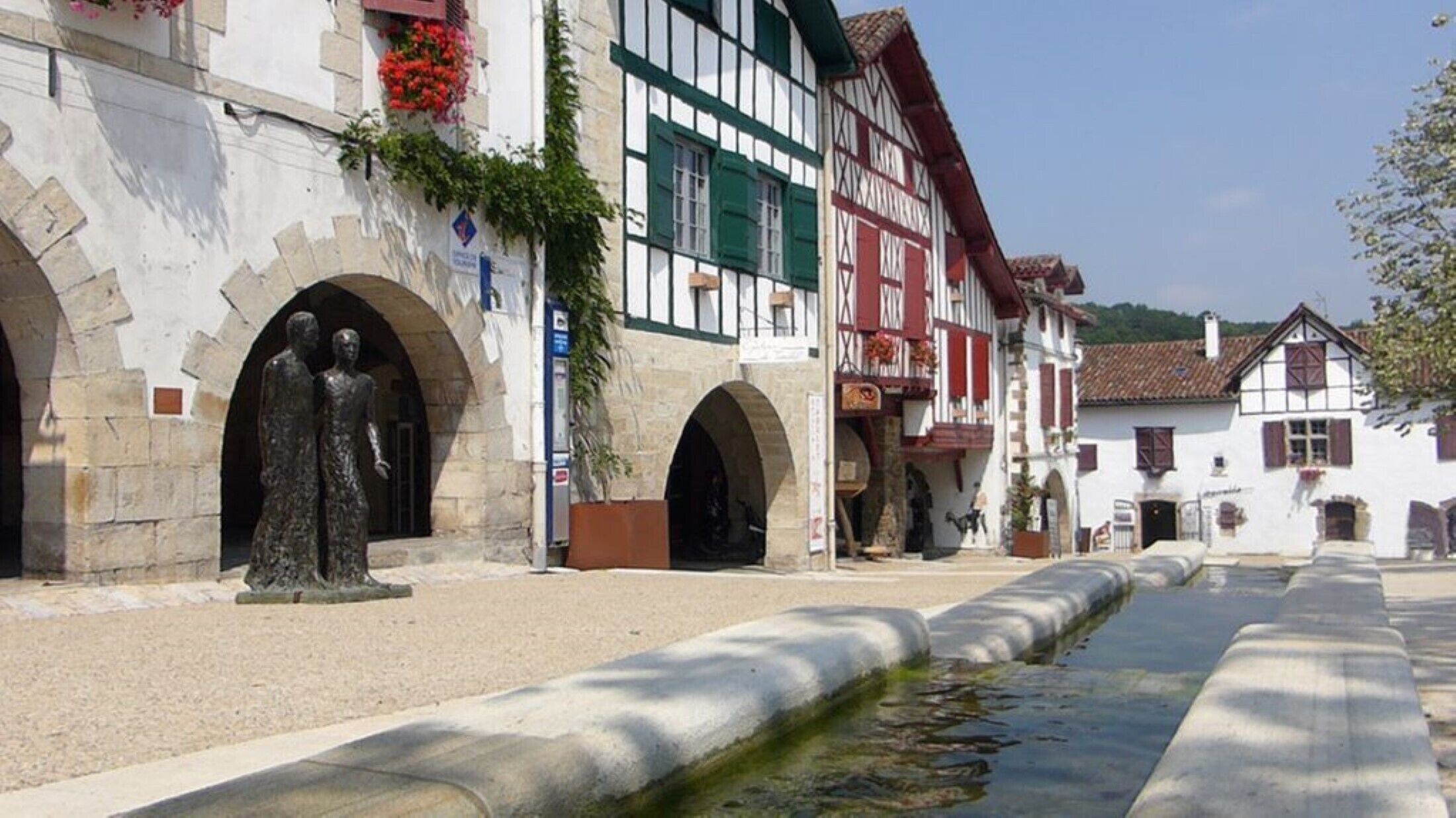A traditional European town square with half-timbered buildings, a statue, and a narrow water channel.