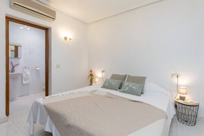 Une chambre d’hôtel avec un lit, des lampes de chevet, une table de nuit, un miroir, une salle de bain visible à travers une porte ouverte et un climatiseur fixé au mur.