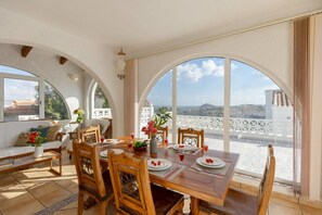 Zona pranzo con tavolo imbandito per quattro persone, ampie finestre ad arco e vista sul paesaggio costiero.