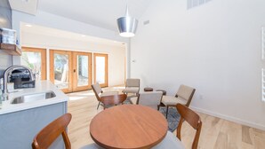 Una cocina moderna con un lavamanos y un horno empotrado, una zona de comedor con una mesa redonda de madera y sillas, y un amplio ventanal corredero que da a un espacio exterior.
