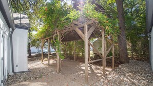 Un espacio al aire libre cubierto con una estructura de pérgola de madera, rodeado de árboles y grava.