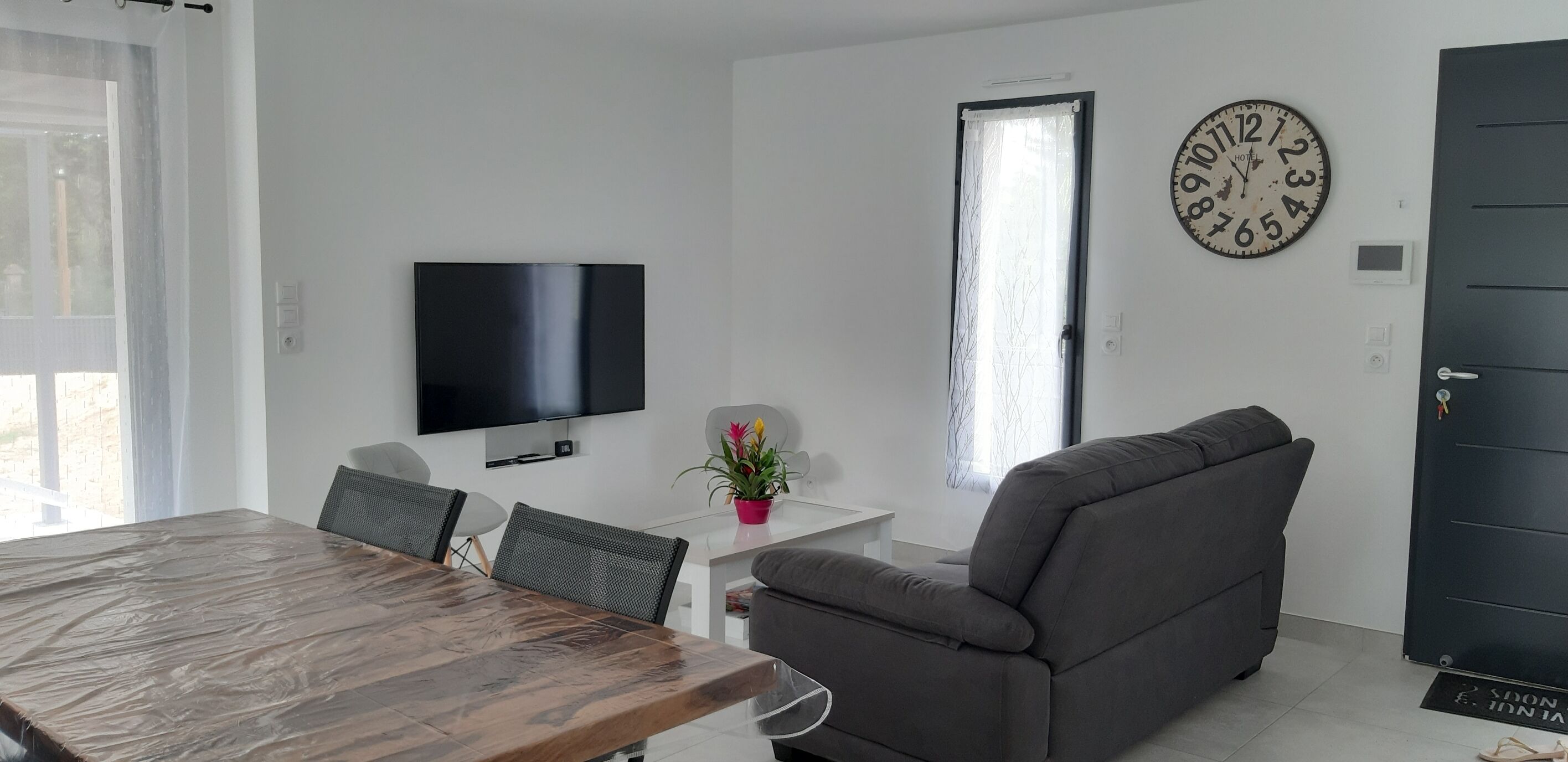 Una sala de estar moderna con una mesa de comedor de madera, un sofá gris, un televisor de pantalla plana y un gran reloj de pared.