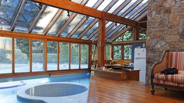 Une piscine intérieure spacieuse, dotée d’une terrasse en bois et de murs en pierre, d’une cheminée en pierre et d’un coin salon confortable agrémenté d’un coussin à rayures.
