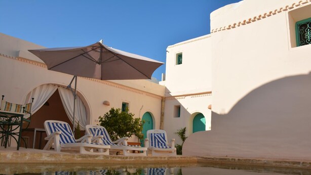 A poolside area with white buildings, blue-striped lounge chairs, and a large umbrella. - Dar Lily (Erriadh)