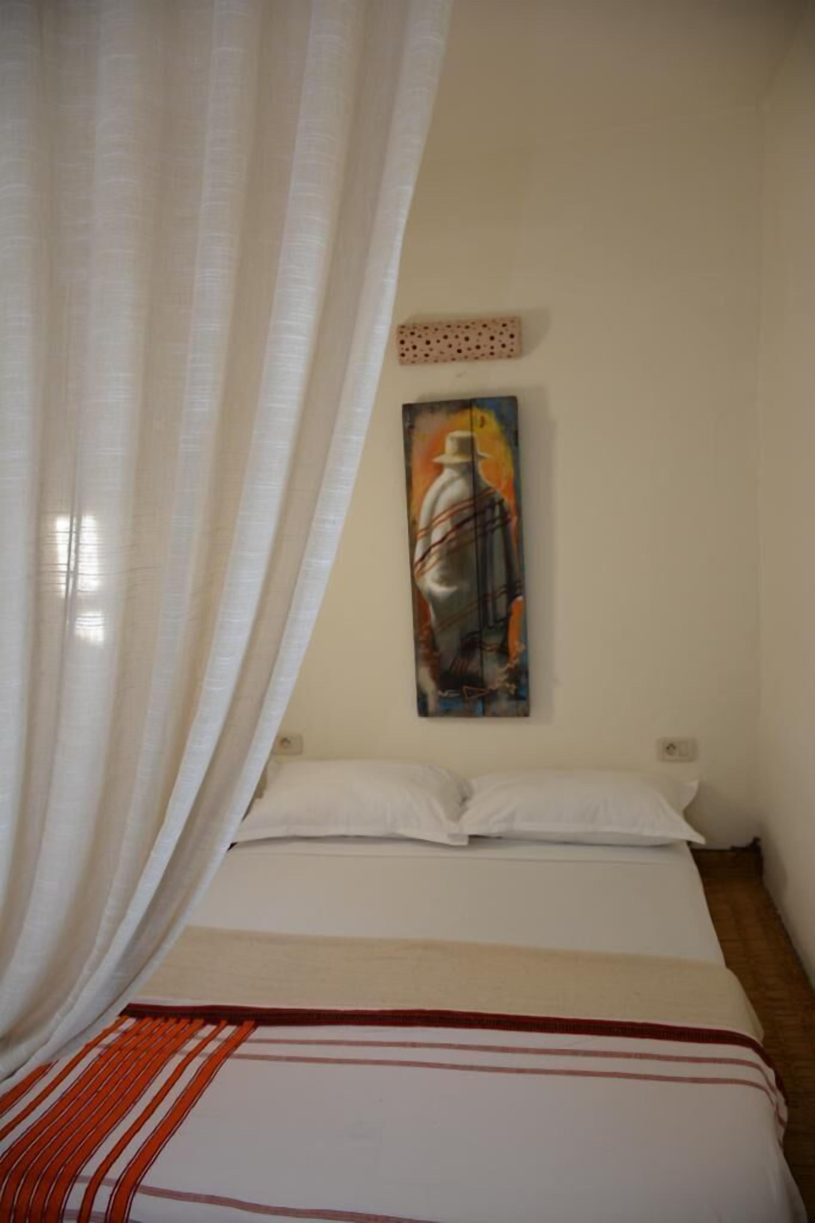 A bedroom with a bed, white curtains, a painting on the wall, and a red-striped blanket.