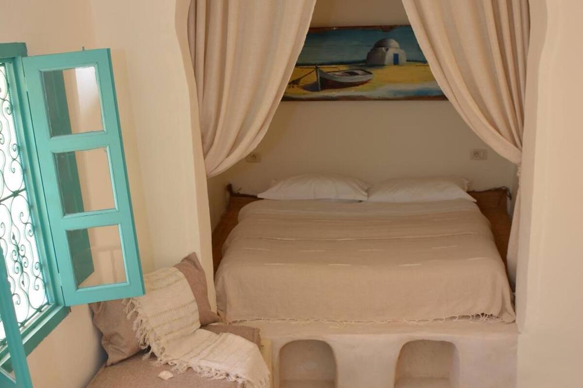 A cozy bedroom with a bed under a canopy, a small table with a cushion, and a patterned rug.