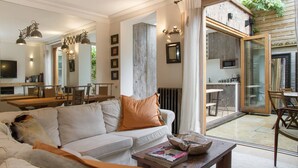 A modern living room with a sofa, coffee table, and dining area. There is a large window with a view of the outdoors. - Retreat to a Stylish Flat in Chelsea (London)
