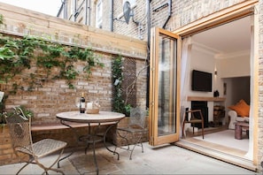 Une terrasse avec une table ronde et des chaises, un mur en briques et une porte-fenêtre coulissante donnant sur une pièce intérieure équipée d’une cheminée et d’un téléviseur.