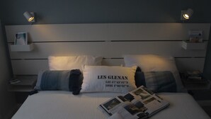 Una cama bien hecha con edredón blanco, dos almohadas y una revista sobre la cama. Hay dos mesitas de noche con lámparas y un pequeño estante en la pared.