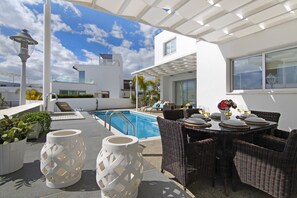 Área de piscina en la azotea con una mesa de comedor para seis personas, rodeada de sillas de mimbre y una pérgola blanca.
