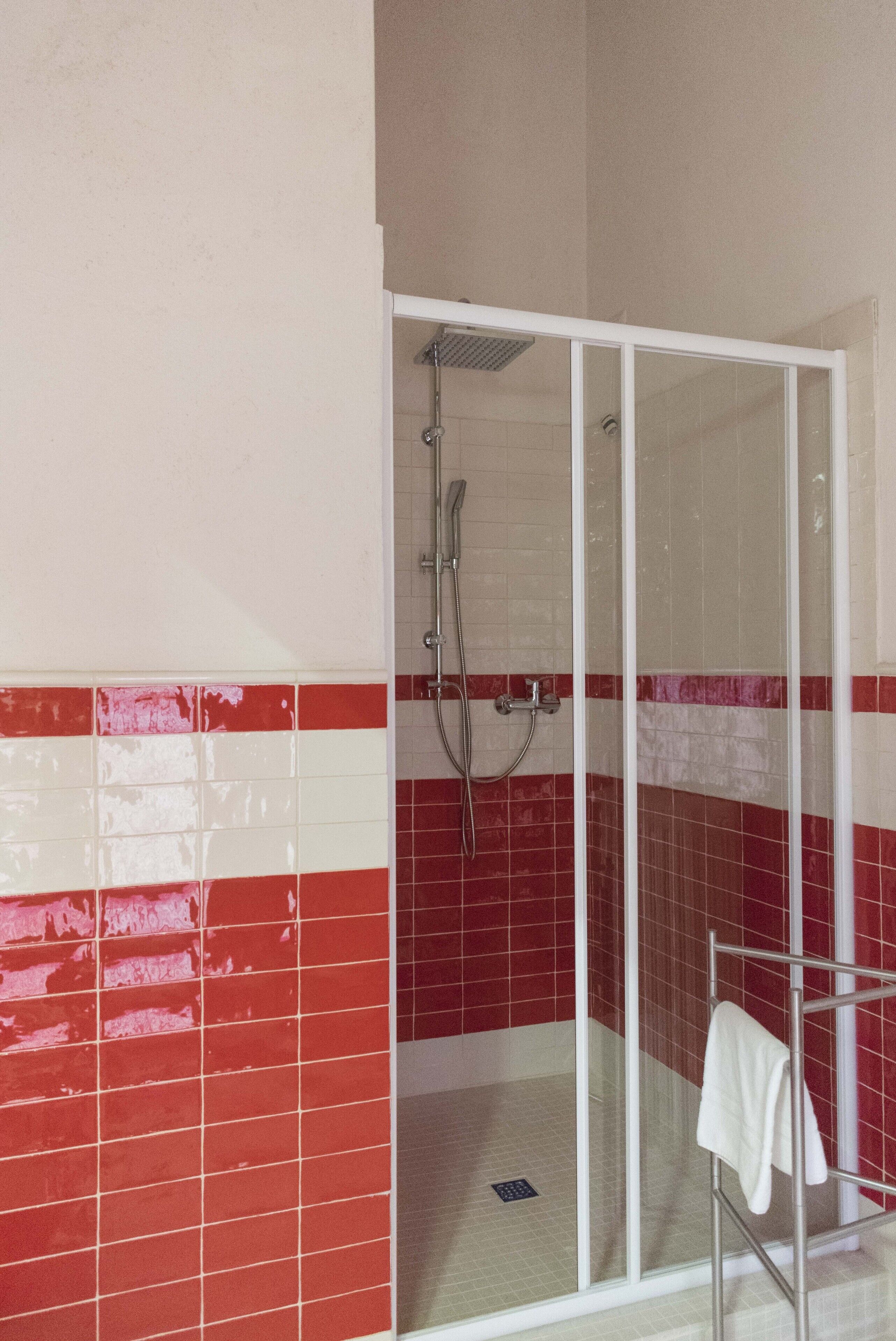 Un área de ducha con puerta de vidrio, paredes con azulejos blancos y rojos, y una toalla blanca en un perchero metálico.