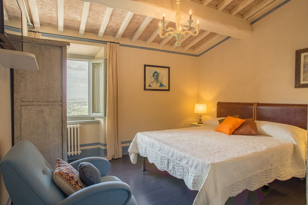 A bedroom with a large bed, a chair, a window with curtains, a chandelier, and a painting on the wall.