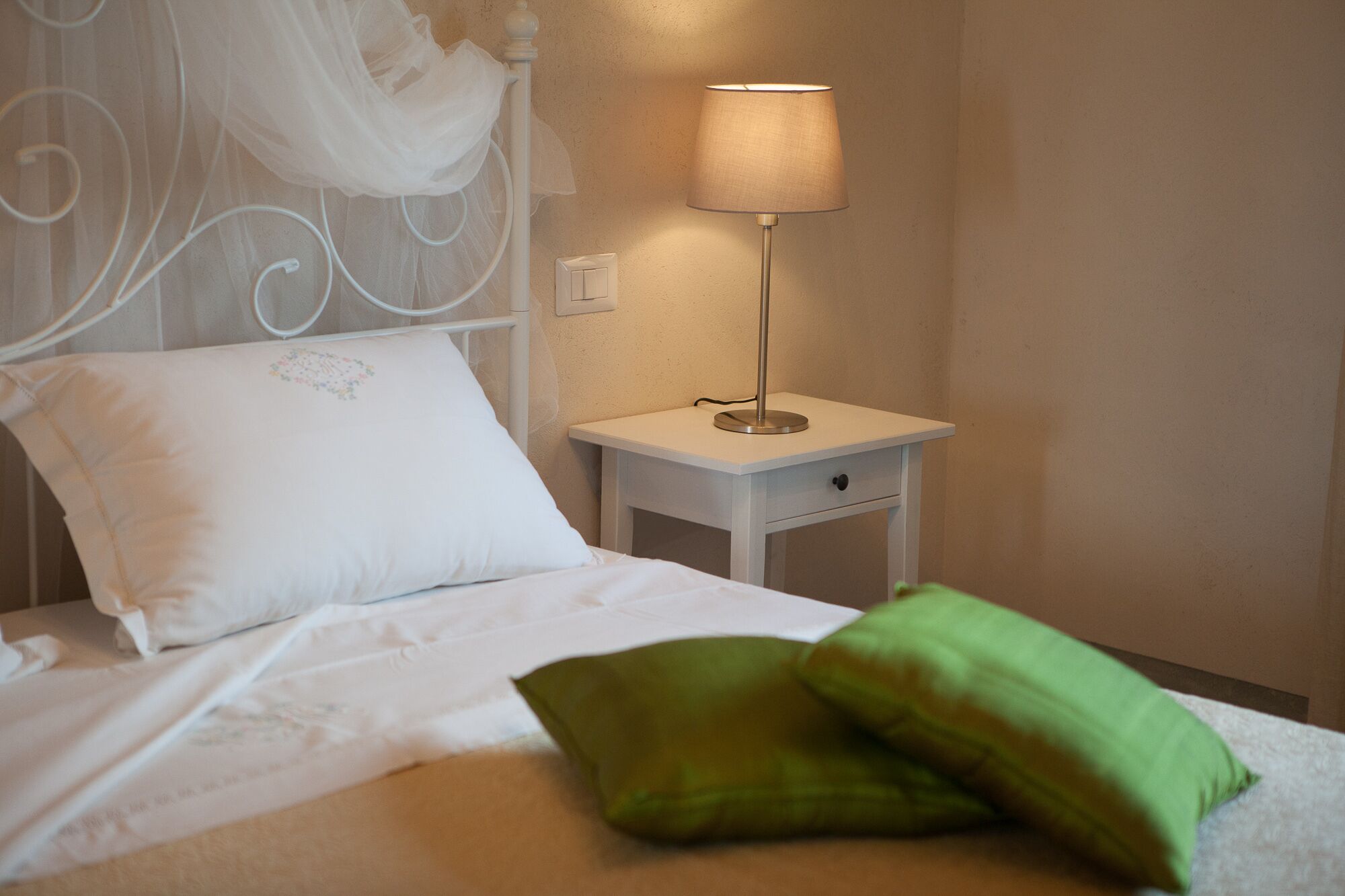 A neatly made bed with white and green pillows, a white bedside table with a lamp, and a wall-mounted switch.