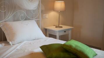A neatly made bed with white and green pillows, a white bedside table with a lamp, and a wall-mounted switch.