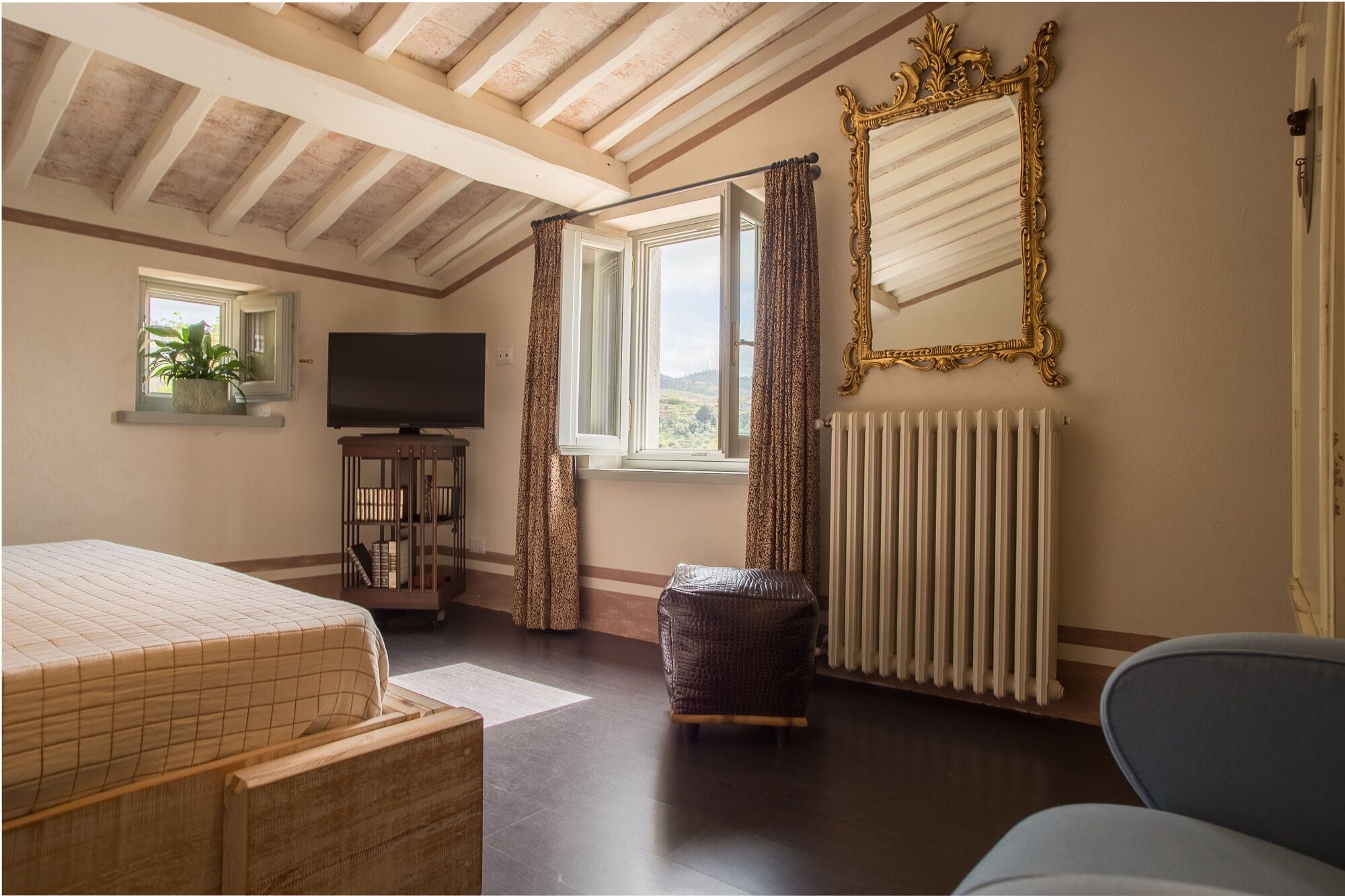 A bedroom with a bed, a TV, a window with curtains, a radiator, and a mirror with an ornate frame.