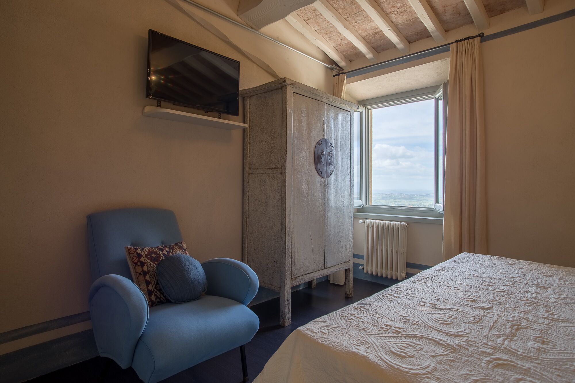 Un dormitorio con una cama, un sillón azul, un armario, un televisor y una ventana con vistas.
