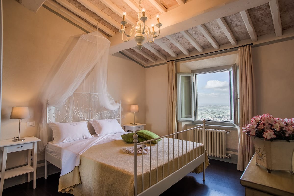 A bedroom with a four-poster bed, a chandelier, a view of the sea through a window, and a radiator.