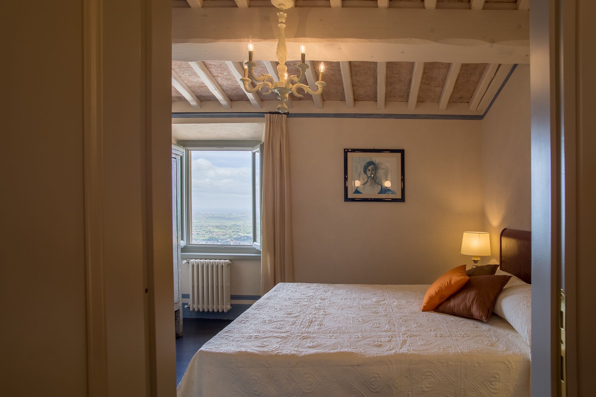Une chambre à coucher avec un grand lit, un lustre, un tableau encadré au mur, une fenêtre avec des rideaux et un radiateur.