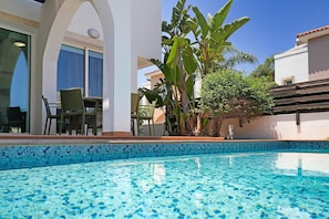 A swimming pool with clear water, surrounded by a patio area with chairs and tables, and lush greenery.