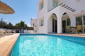 A swimming pool with clear blue water, surrounded by a white building with arched doorways and a covered patio area with lounge chairs. - MADISON - 3Bed Villa with private pool within walking distance to Pernera Resort Center (Paralimni)