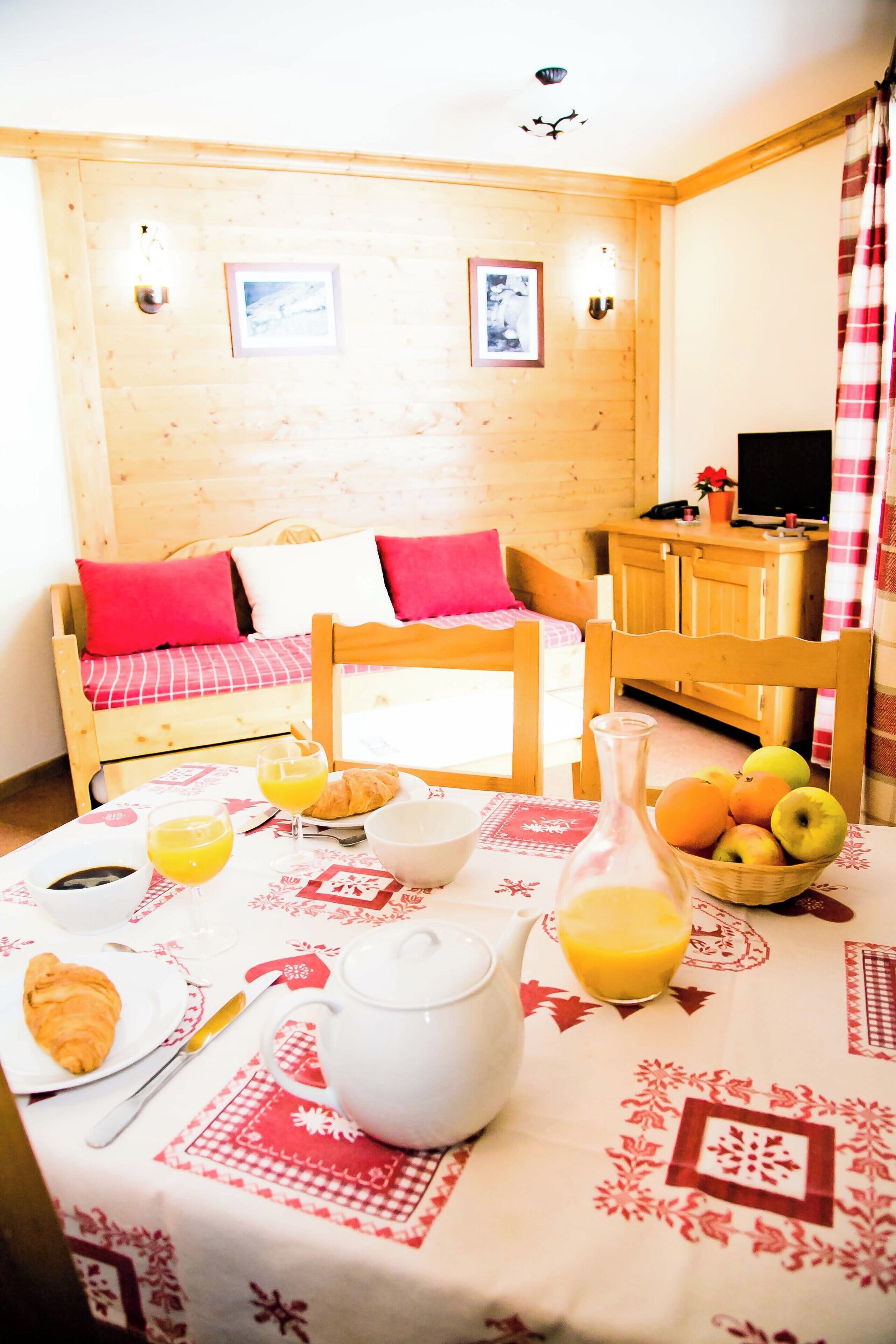 A cozy room with a wooden wall, a bed with red and white bedding, a dining table with breakfast items, and a television on a wooden cabinet.