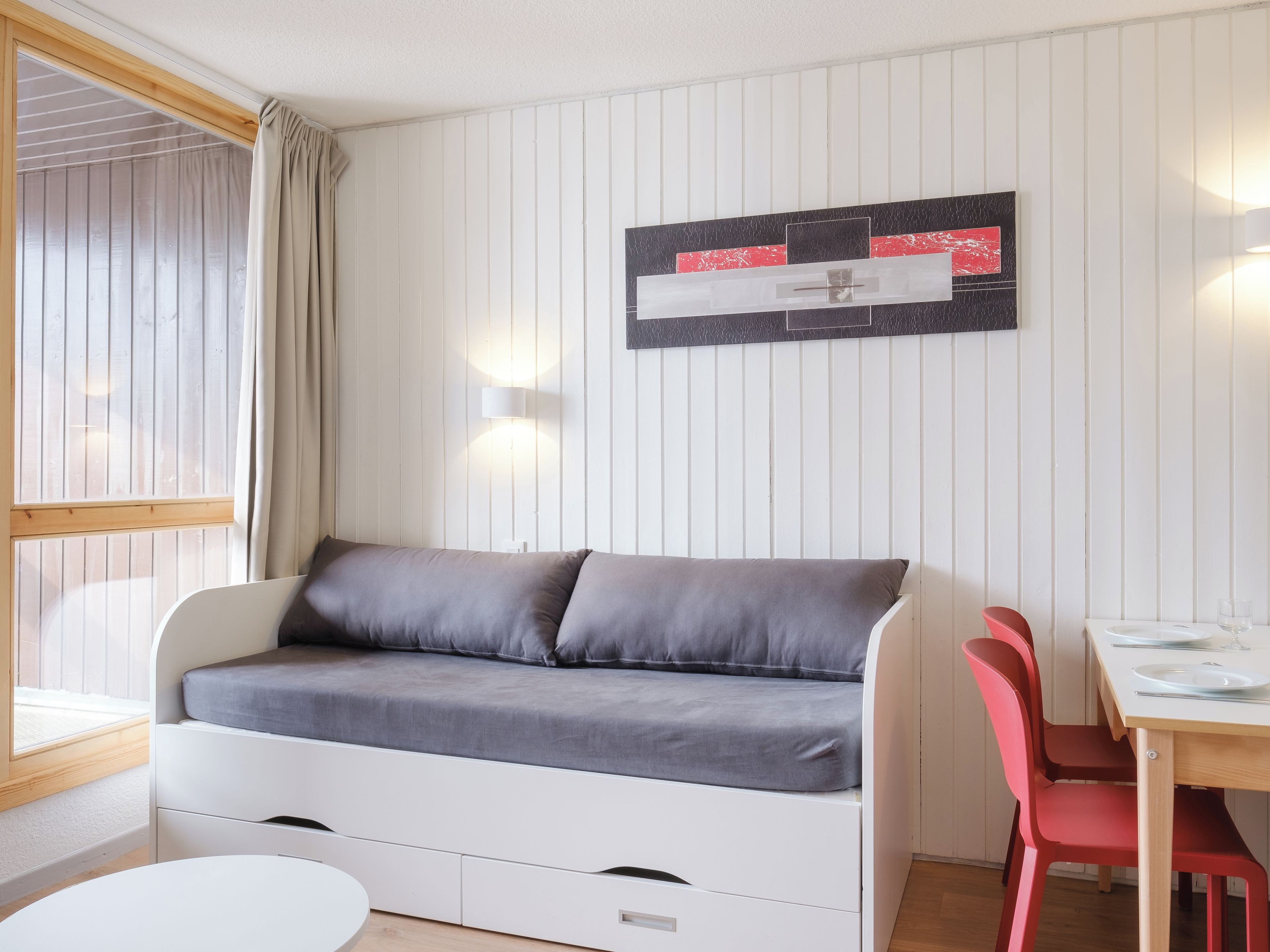 Une pièce avec un canapé blanc, une table à manger entourée de chaises rouges et un luminaire fixé au mur.
