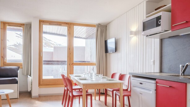 Uma cozinha moderna com micro-ondas, pia e lava-louças. Há uma área de jantar com mesa e cadeiras, e uma televisão fixada na parede.