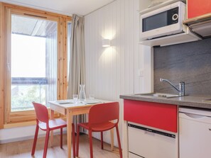 Une cuisine compacte équipée d’un four à micro-ondes, d’un évier et d’un lave-vaisselle. On y trouve une table à manger avec deux chaises et une fenêtre avec des rideaux.