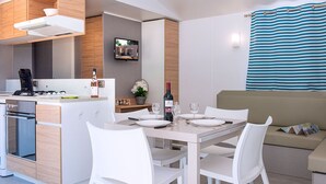 A modern kitchen with white cabinetry, a dining area with a table set for four, and a living area with a sofa and a striped curtain. - Camping Sunissim Le Domaine Les Mûriers (Vendres)