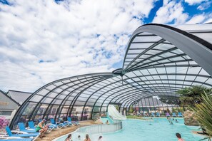 A covered swimming pool with a glass ceiling, lounge chairs, and a water slide. - Camping Sunissim Emeraude (Saint-Briac-sur-Mer)