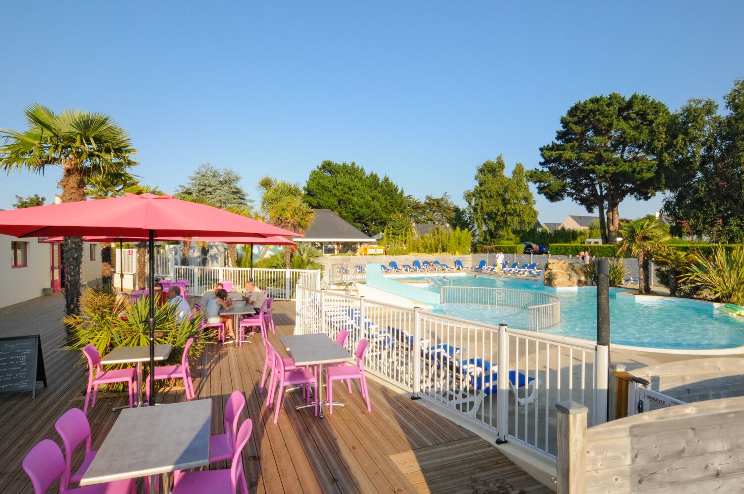 Un espace aménagé au bord de la piscine, avec une terrasse en bois, des chaises roses et des tables placées sous un parasol rouge.