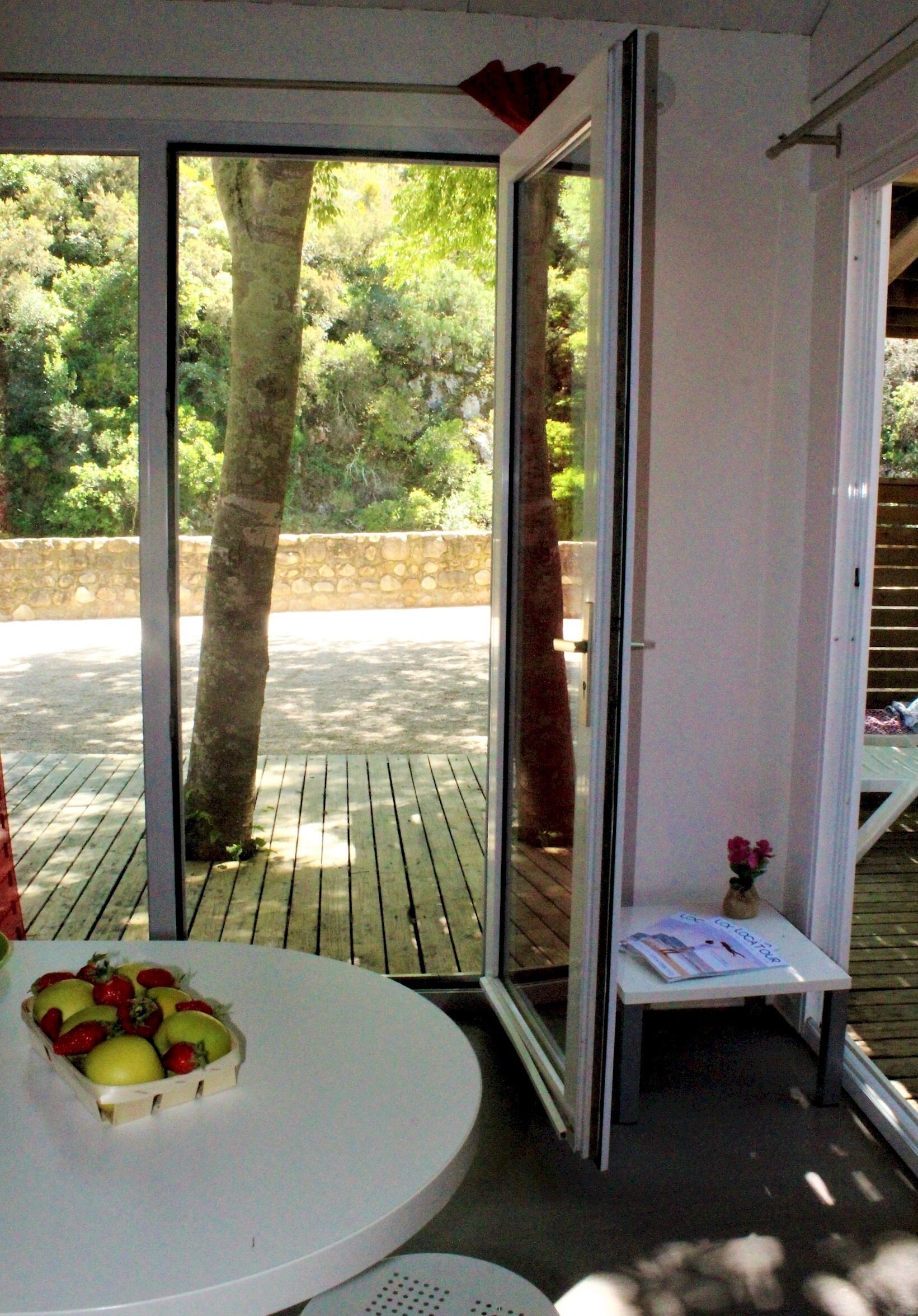 Un patio couvert avec une table, une petite étagère sur laquelle est posé un vase, et une vue sur un arbre et la verdure à l’extérieur.