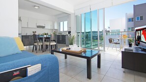 Una sala de estar moderna con un sofá, una mesa de centro y un televisor. Se observa una cocina al fondo con una zona de comedor.