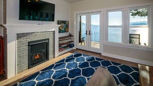 Un salon doté d’une cheminée, d’un téléviseur, offrant une vue sur la mer grâce à de grandes fenêtres, et agrémenté d’un tapis bleu à motifs.