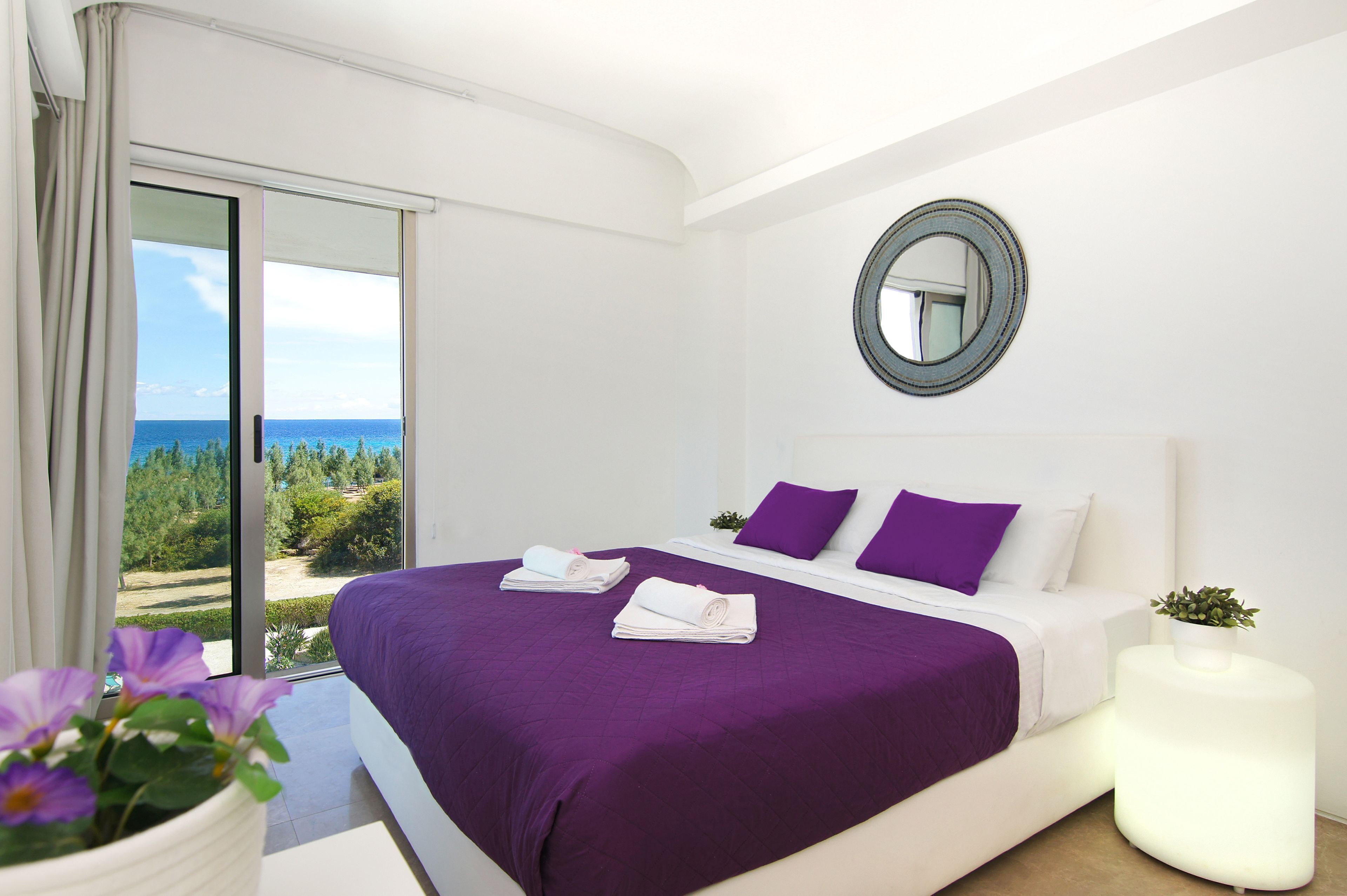 A bedroom with a bed, a round mirror, a potted plant, and a view of the sea.