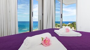 Una habitación con una colcha morada, toallas blancas con flores rosadas y vistas al océano a través de amplios ventanales.