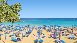 A beach with numerous sun loungers and umbrellas, a clear blue sky, and the ocean in the background.