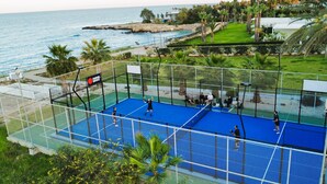 A tennis court with a net, surrounded by a fence, and overlooking a coastal area with palm trees and a view of the sea.