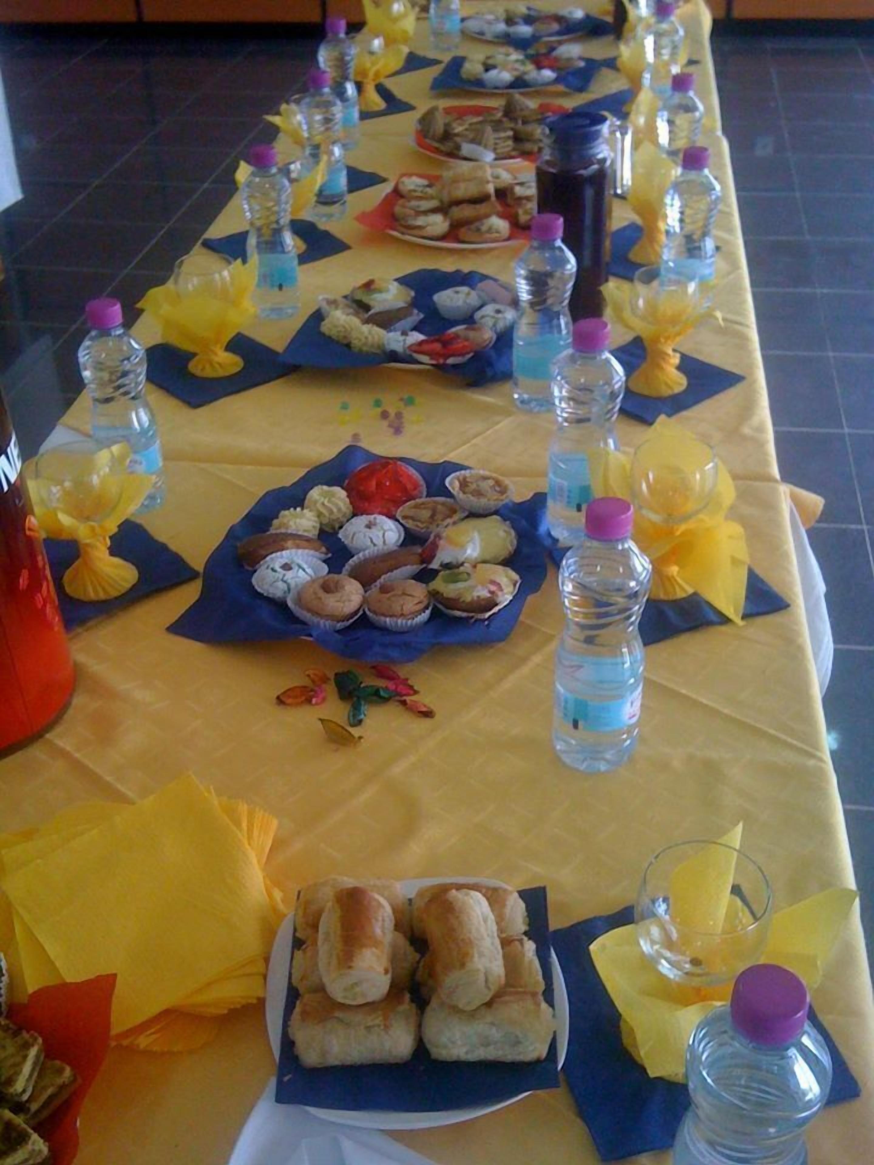 Une longue table, recouverte d’une nappe jaune, sur laquelle sont disposés divers aliments et des bouteilles d’eau.