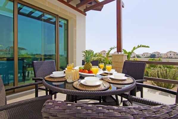 A dining table set with plates, cutlery, and a fruit bowl on a balcony with wicker chairs and a glass table.