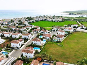 Vue aérienne d’un quartier résidentiel situé sur la côte, avec des bâtiments blancs, un grand espace vert central et une piscine.