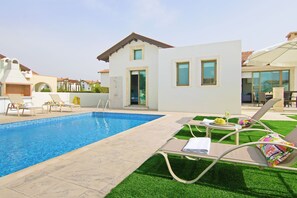 Une piscine entourée de chaises longues et d’une petite table où sont servis des boissons, le tout bordé d’une pelouse bien entretenue et d’un bâtiment moderne doté de grandes fenêtres.
