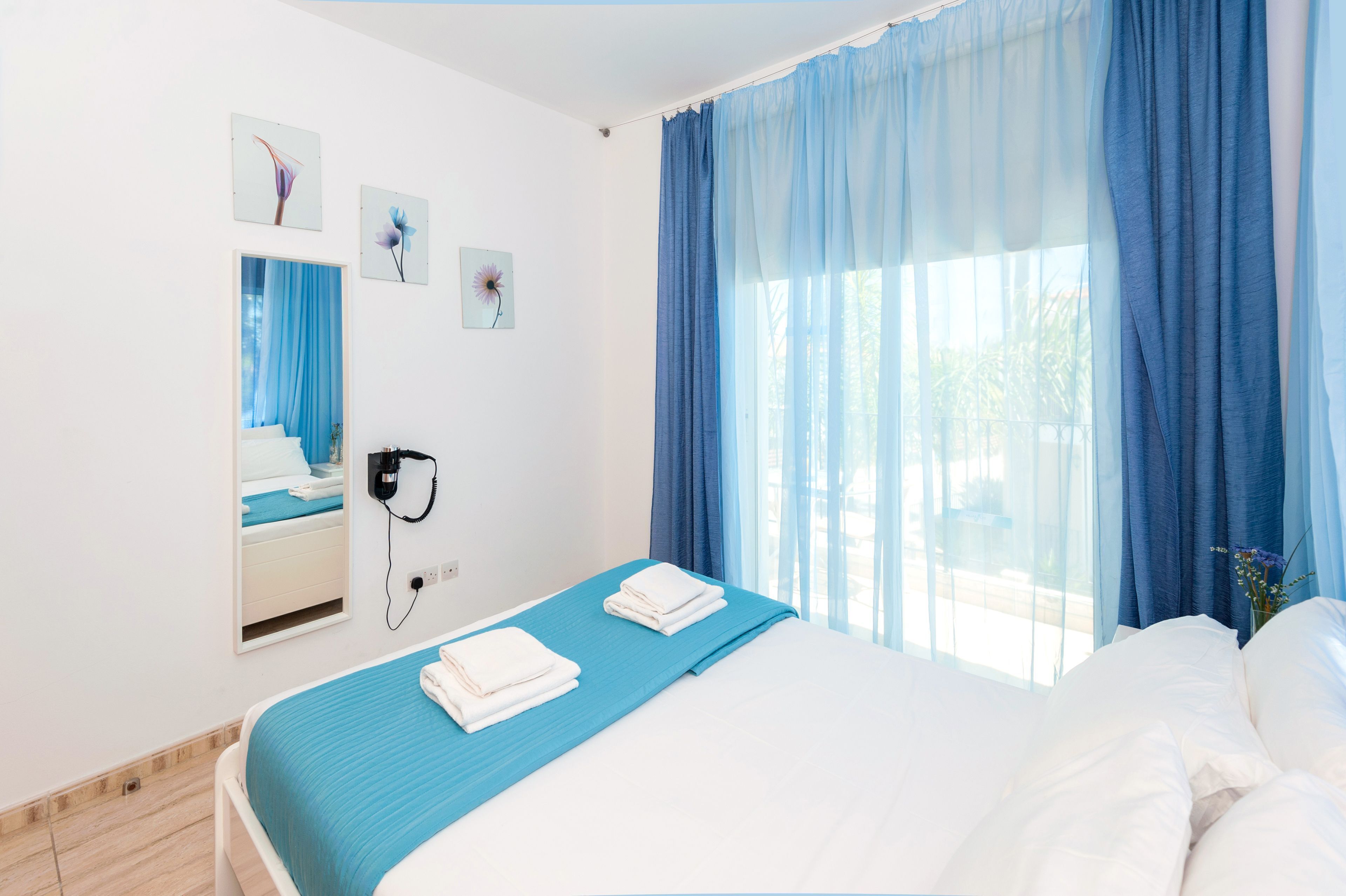 Ein Hotelzimmer mit Bett, Spiegel, Fenster mit blauen Vorhängen und Bildern an der Wand.