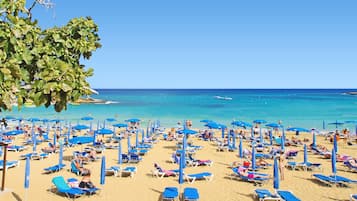 A beach with numerous sun loungers and umbrellas, a clear blue sky, and the ocean in the background.