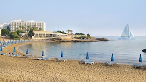 Une plage bordée de rangées de chaises longues et de parasols, un voilier sur l’eau et un hôtel en arrière-plan.