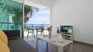 A modern living room with a sofa, a coffee table, and a TV on a stand. There is a balcony with a view of the sea. - NIOVI Sea View Aprt - Coralli Beachfront Resort (Paralimni)