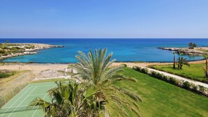A coastal view with a tennis court, palm trees, and a clear blue sea. - NIOVI Sea View Aprt - Coralli Beachfront Resort (Paralimni)